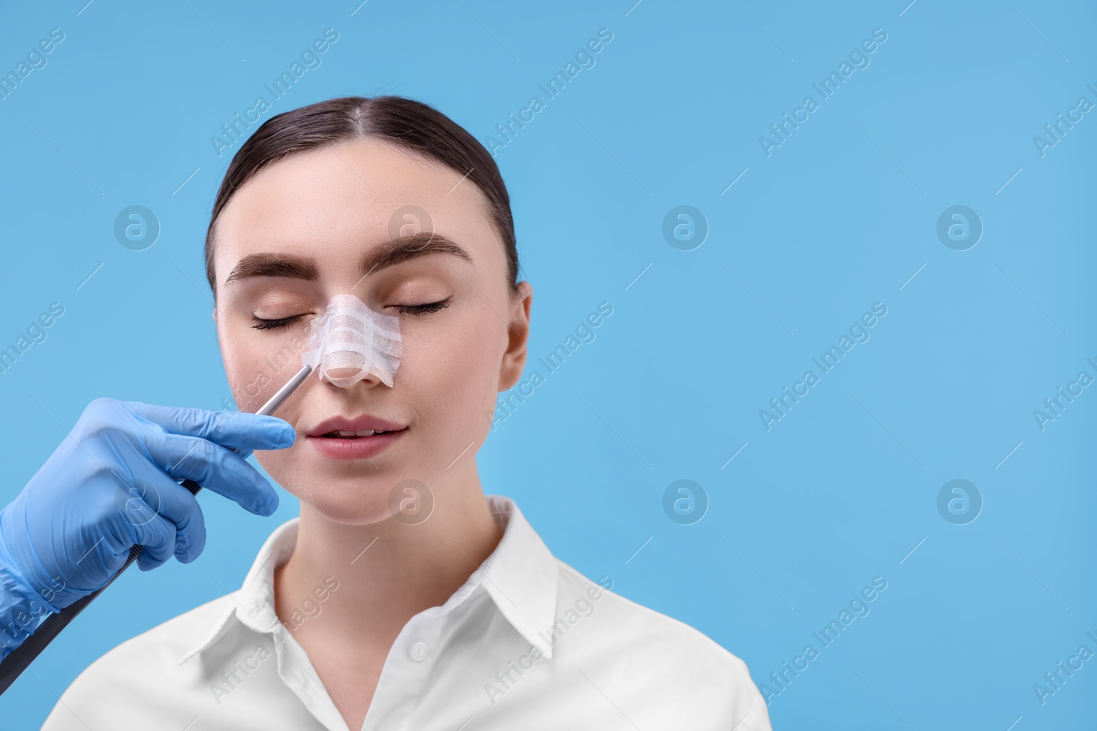 Photo of Doctor removing medical bandage from patient's nose after plastic surgery operation on light blue background, closeup. Space for text