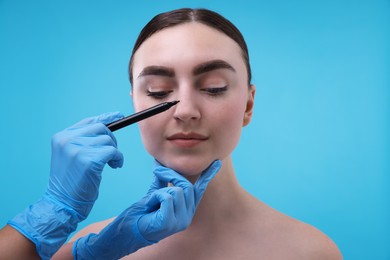 Photo of Doctor drawing marks on patient's nose for plastic surgery operation against light blue background, closeup