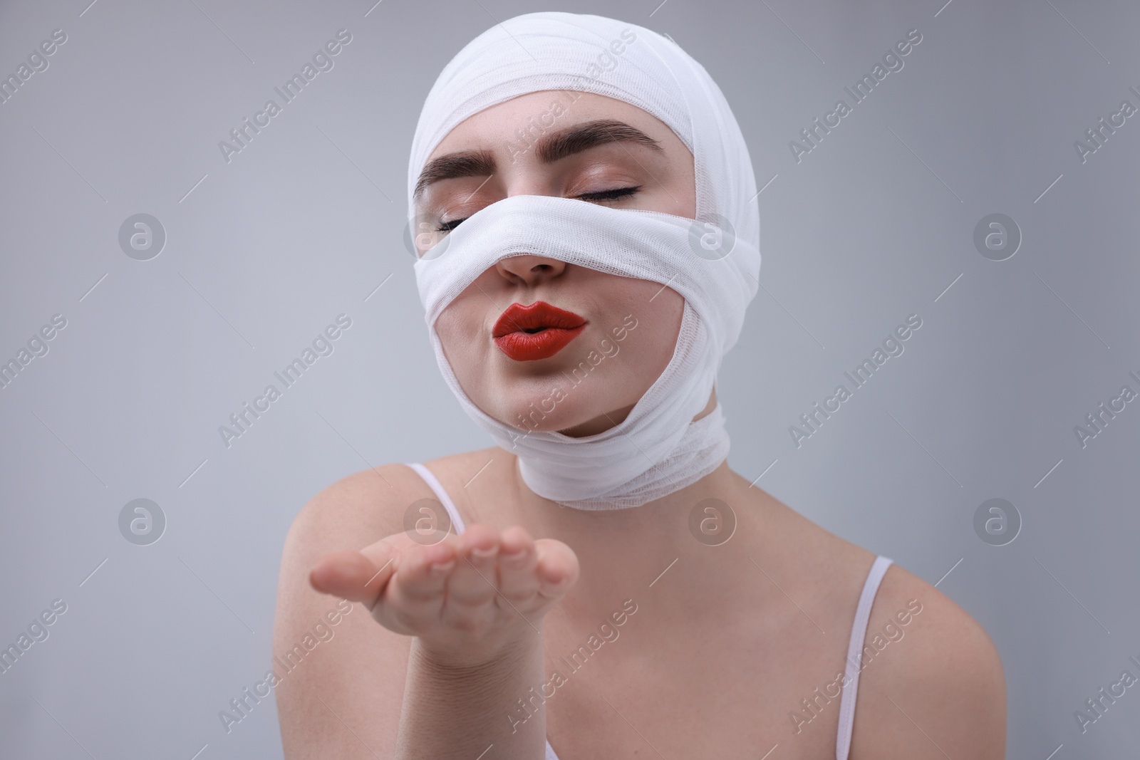 Photo of Woman with nose wrapped in medical bandage after plastic surgery operation blowing kiss on grey background