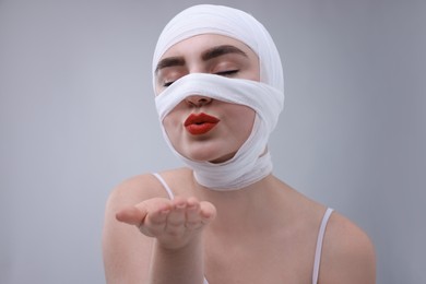 Woman with nose wrapped in medical bandage after plastic surgery operation blowing kiss on grey background