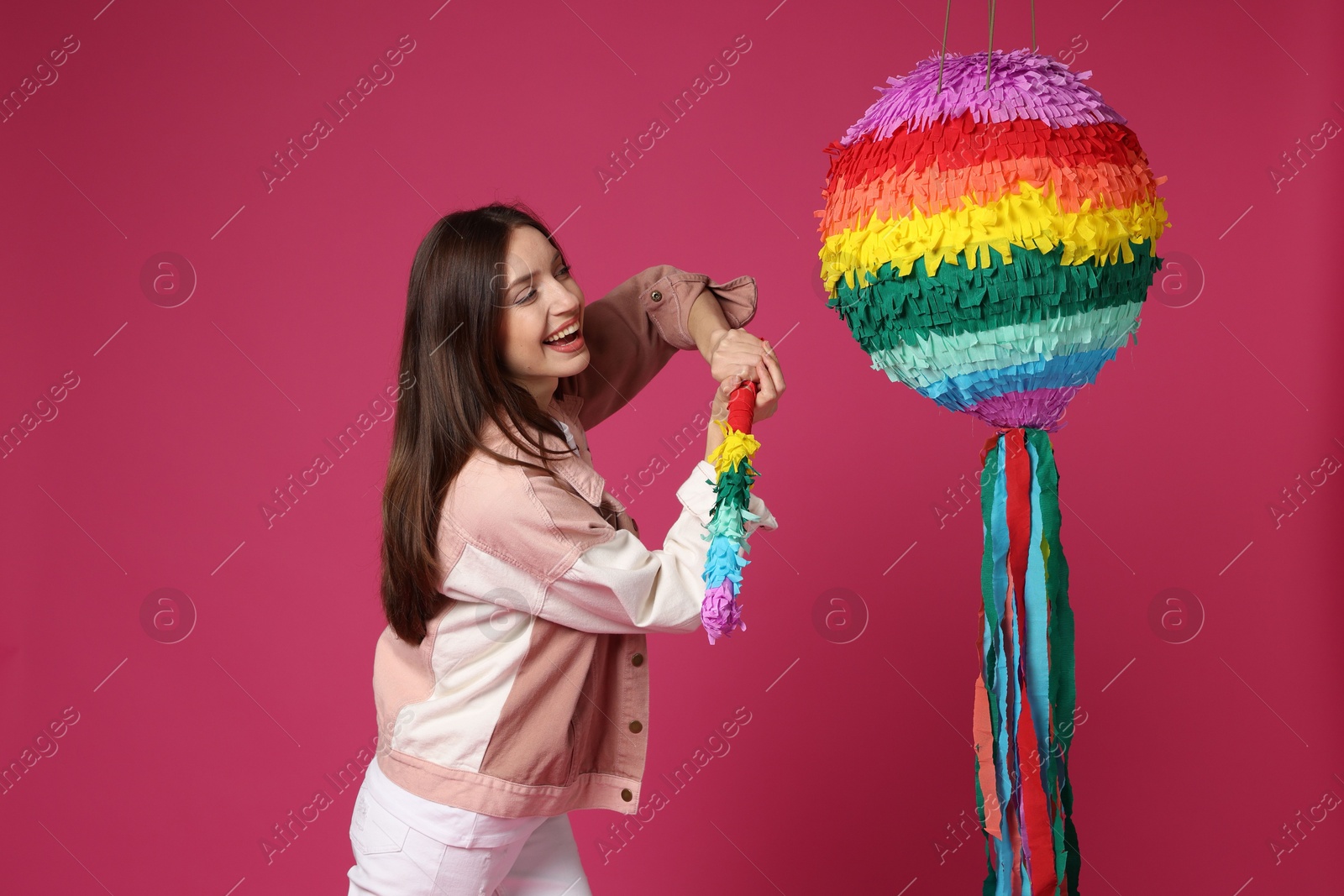 Photo of Happy woman breaking colorful pinata with stick on pink background