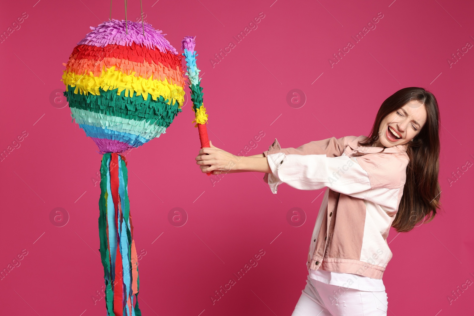 Photo of Happy woman breaking colorful pinata with stick on pink background