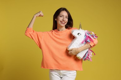 Photo of Happy woman with unicorn shaped pinata showing her strength on yellow background