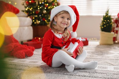 Photo of Little girl in Santa hat with gift box sitting on floor at home