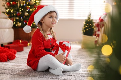 Photo of Little girl in Santa hat with gift box sitting on floor at home