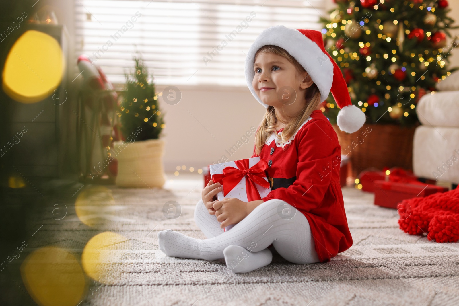 Photo of Little girl in Santa hat with gift box sitting on floor at home