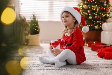 Photo of Little girl in Santa hat with gift box sitting on floor at home