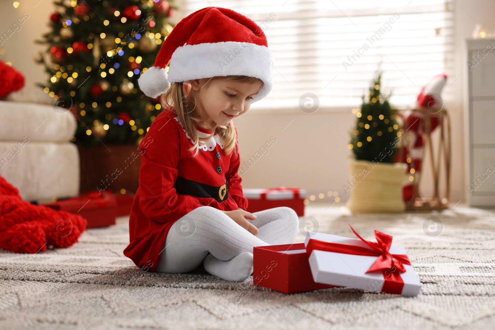 Photo of Little girl in Santa hat with gift box sitting on floor at home