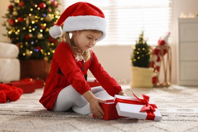 Photo of Little girl in Santa hat with gift box sitting on floor at home