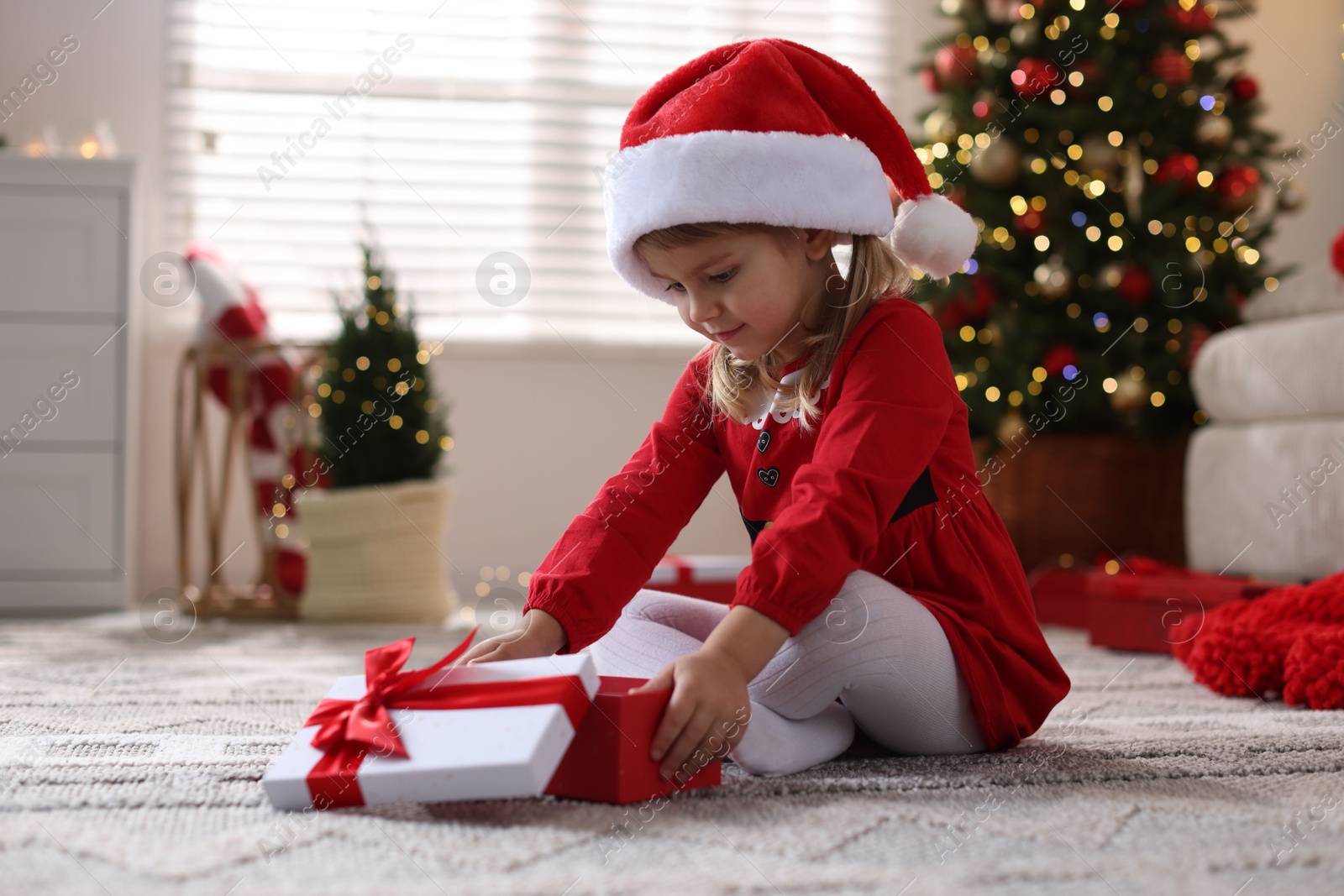 Photo of Little girl in Santa hat with gift box sitting on floor at home