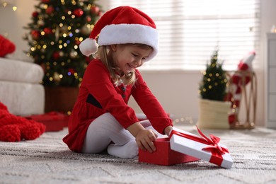 Photo of Little girl in Santa hat with gift box sitting on floor at home