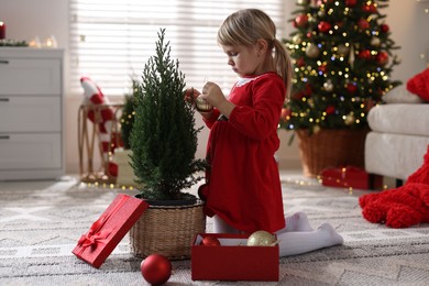 Photo of Little girl in Christmas costume with gift box decorating small tree at home