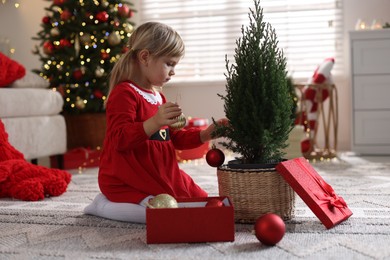 Photo of Little girl in Christmas costume with gift box decorating small tree at home