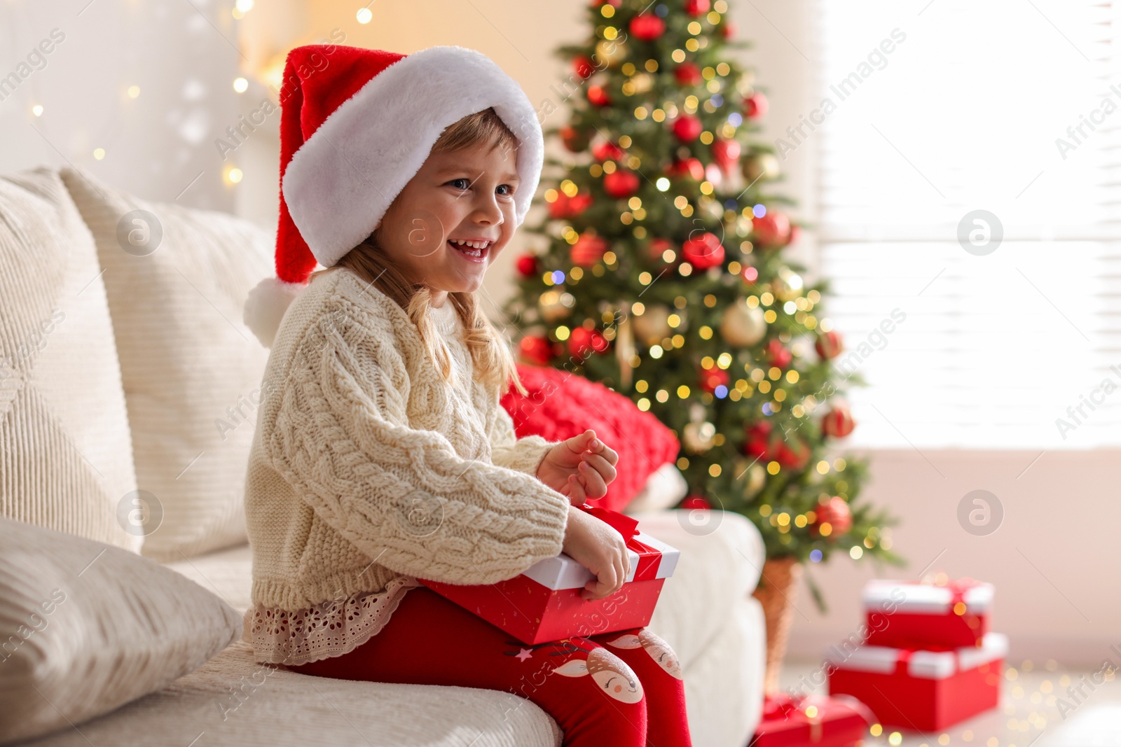 Photo of Little girl in Santa hat sitting on sofa at home