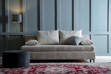 Photo of Comfortable sofa with pillows, lamp and pouf near grey wall indoors