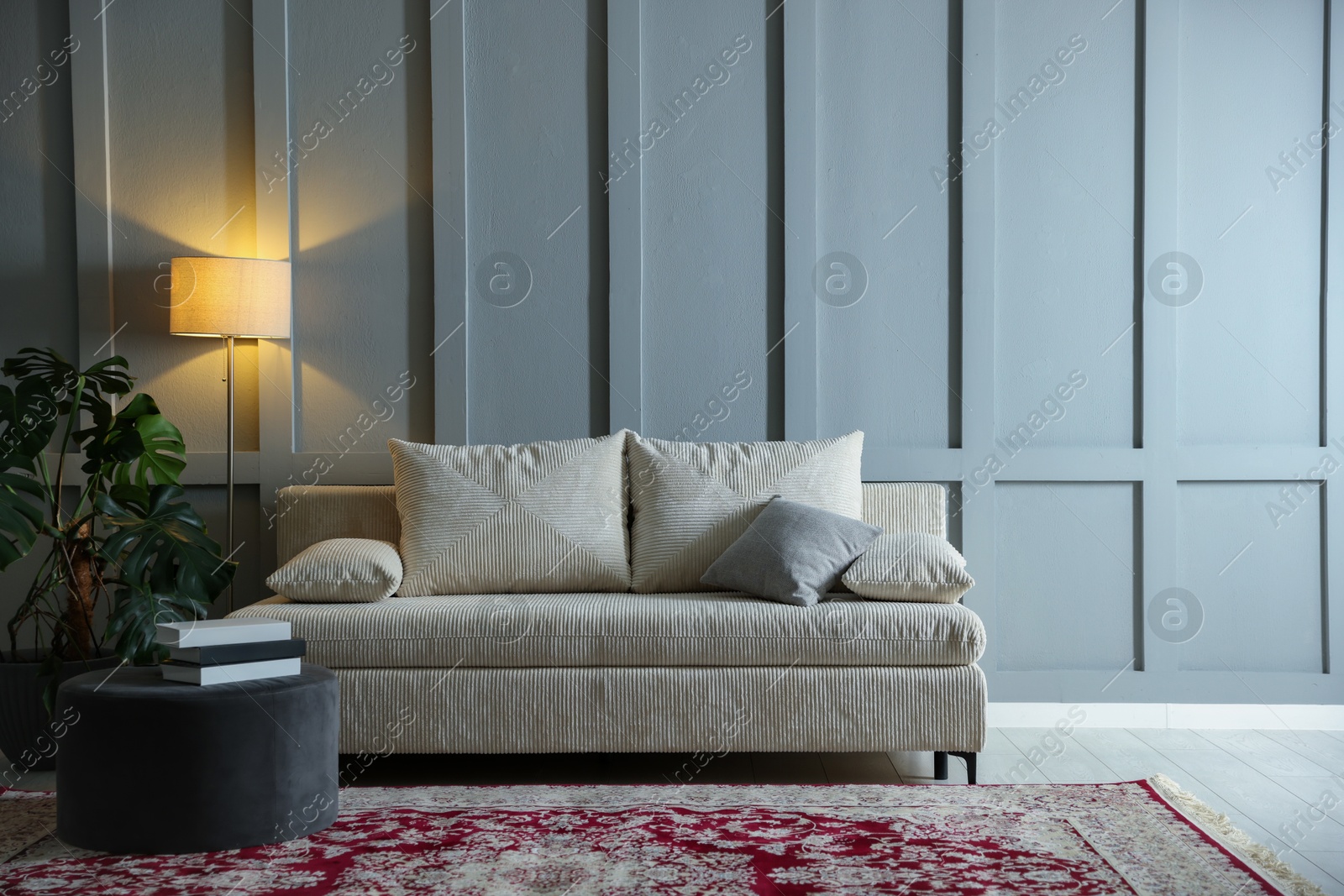 Photo of Comfortable sofa with pillows, lamp, houseplant and pouf near grey wall indoors. Space for text