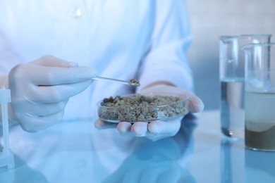 Photo of Laboratory testing. Scientist holding petri dish with sand sample at table indoors, closeup