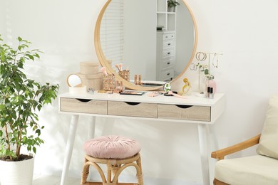 Photo of Makeup room. Dressing table with mirror, different beauty products, stool, armchair and houseplant