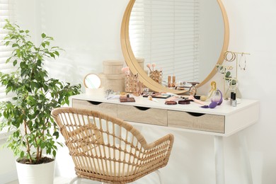 Photo of Makeup room. Dressing table with mirror, different beauty products, jewelry, chair and houseplant indoors