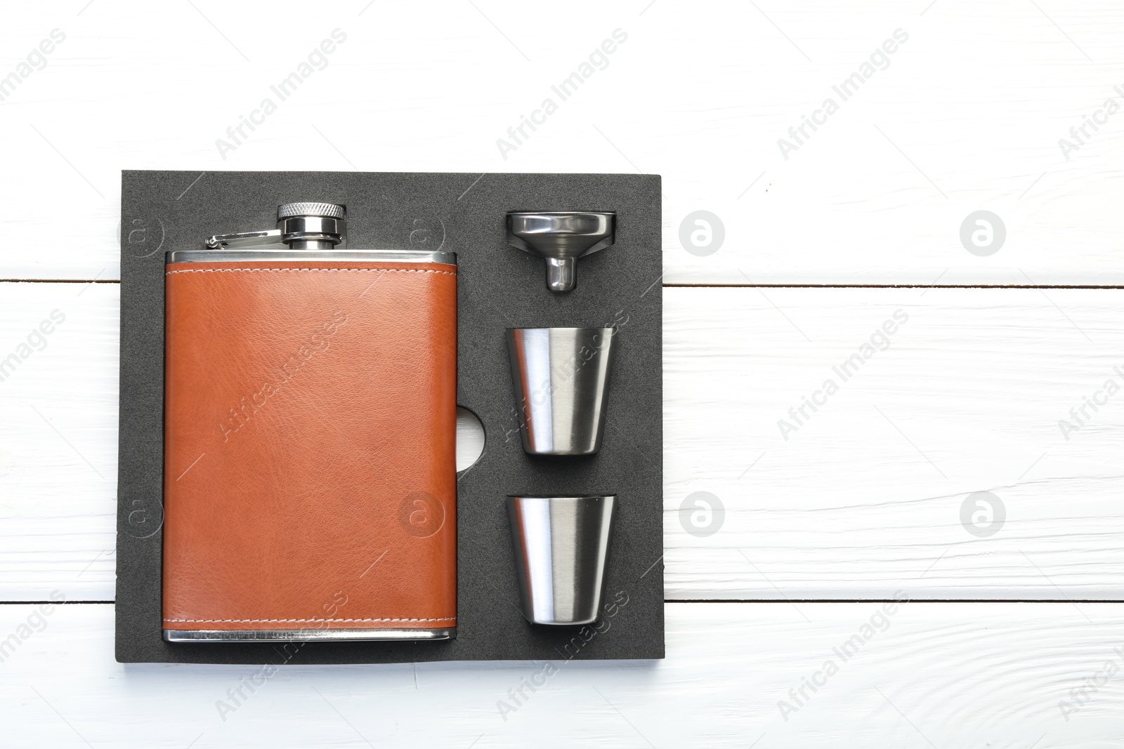 Photo of Hip flask, cups and funnel in package on white wooden table, top view. Space for text