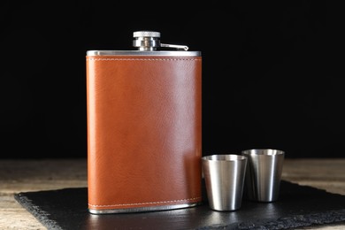 Photo of Hip flask and cups on wooden table against black background