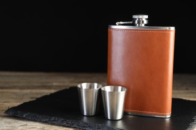 Photo of Hip flask and cups on wooden table against black background, space for text