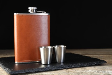 Photo of Hip flask and cups on wooden table against black background, space for text