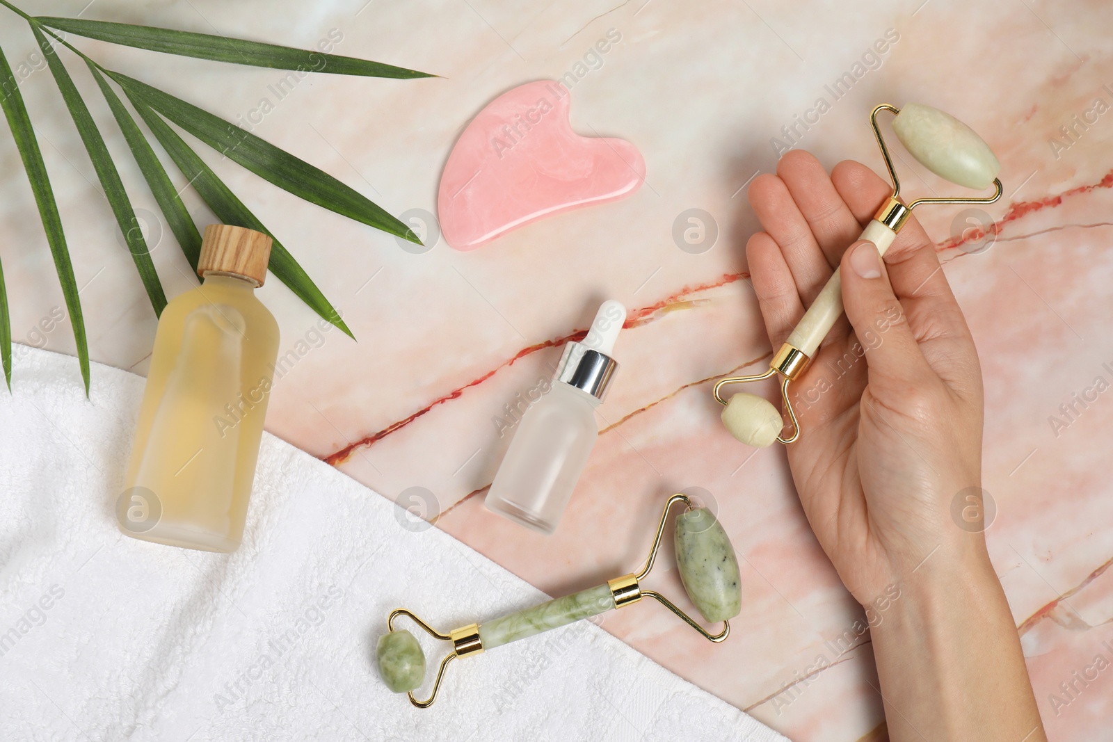 Photo of Woman with face rollers, gua sha tool, cosmetic products, towel and leaf on pink marble background, top view