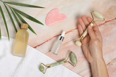 Photo of Woman with face rollers, gua sha tool, cosmetic products, towel and leaf on pink marble background, top view