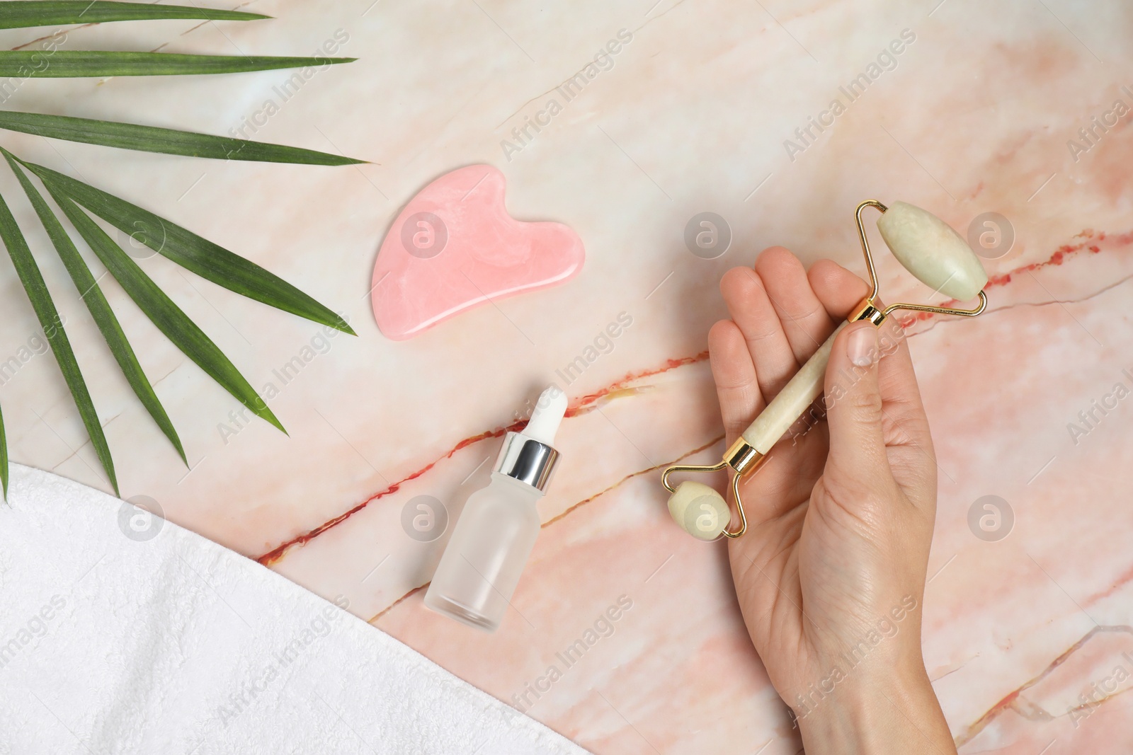 Photo of Woman with face roller, gua sha tool, cosmetic product, towel and leaf on pink marble background, top view