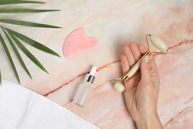 Photo of Woman with face roller, gua sha tool, cosmetic product, towel and leaf on pink marble background, top view