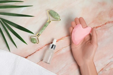 Photo of Woman with gua sha tool, face roller, cosmetic product, towel and leaf on pink marble background, top view