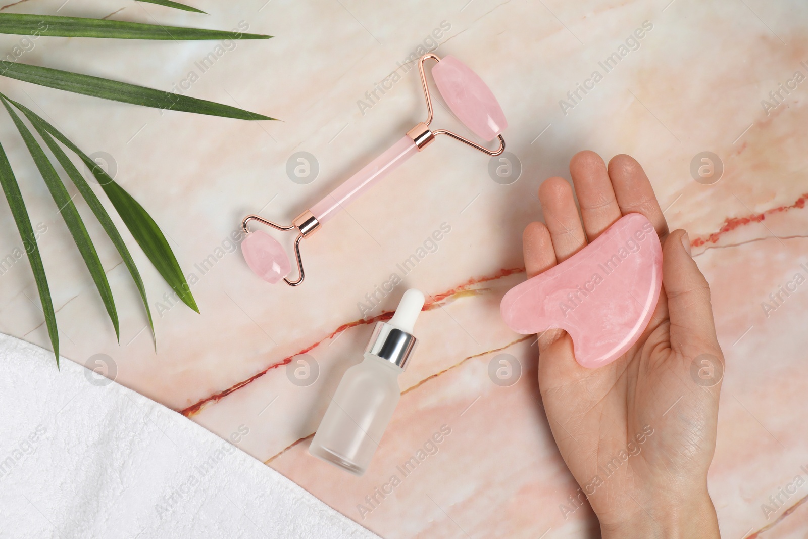 Photo of Woman with gua sha tool, face roller, cosmetic product, towel and leaf on pink marble background, top view