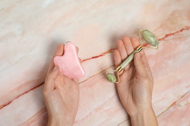 Photo of Woman with face roller and gua sha tool on pink marble background, top view