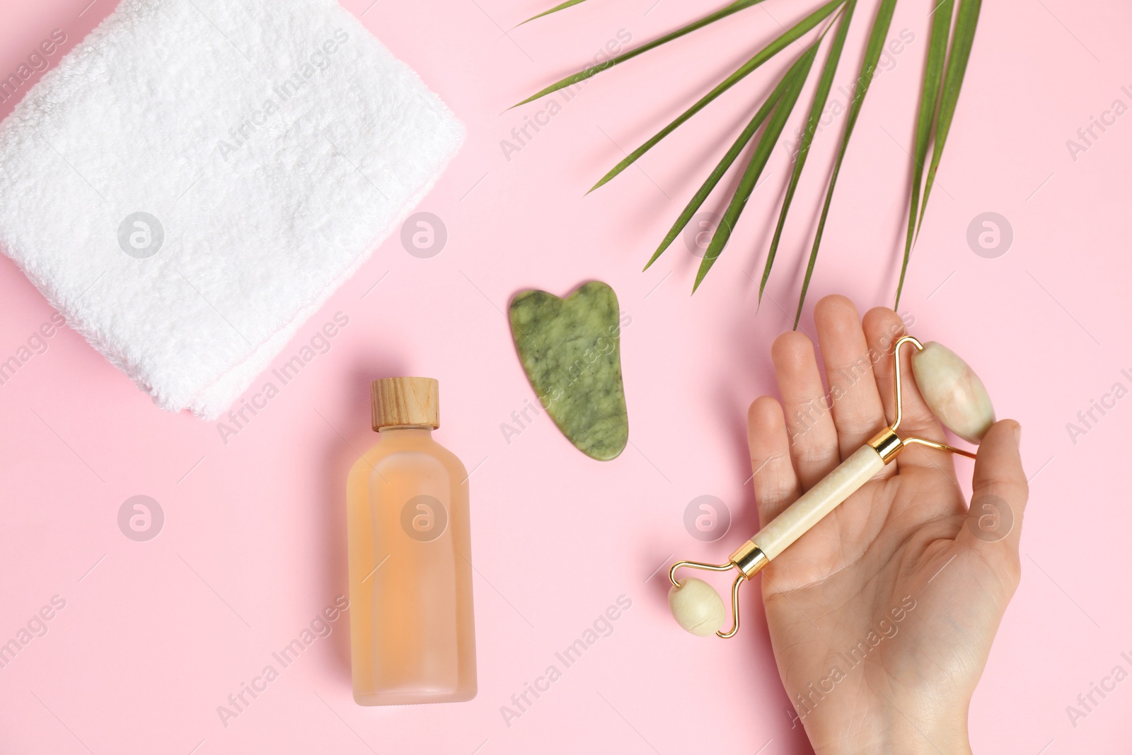 Photo of Woman with face roller, gua sha tool, cosmetic product, towel and leaf on pink background, top view