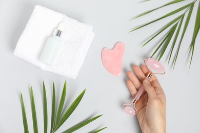 Photo of Woman with face roller, gua sha tool, cosmetic product, towel and leaves on light background, top view