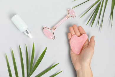 Photo of Woman with gua sha tool, face roller, cosmetic product and leaves on light background, top view