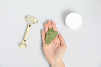 Photo of Woman with gua sha tool, face roller and cosmetic product on light background, top view