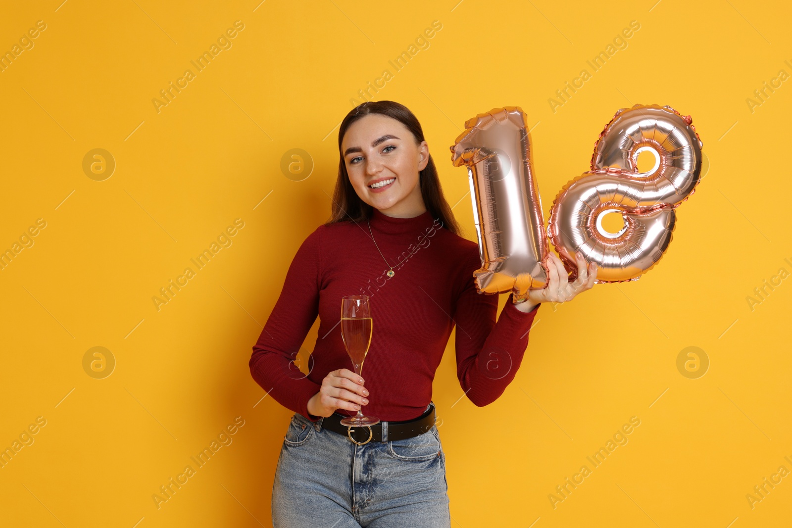 Photo of Coming of age party - 18th birthday. Happy young woman with number shaped balloons and glass of sparkling wine on yellow background