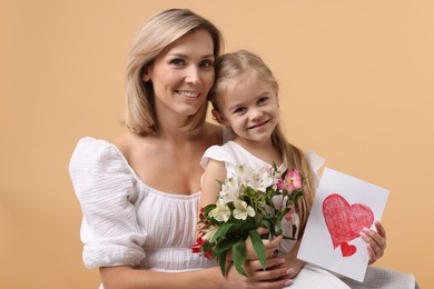 Photo of Happy woman with her daughter, bouquet of alstroemeria flowers and greeting card on beige background. Mother's Day celebration