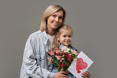 Photo of Happy woman with her daughter, bouquet of alstroemeria flowers and greeting card on gray background. Mother's Day celebration