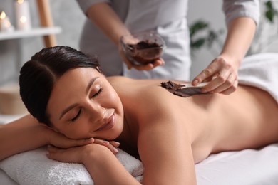 Photo of Chocolate body wrap. Spa worker applying mask onto woman's back in salon, closeup