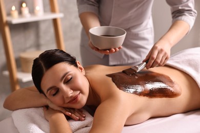 Photo of Chocolate body wrap. Spa worker applying mask onto woman's back in salon, closeup