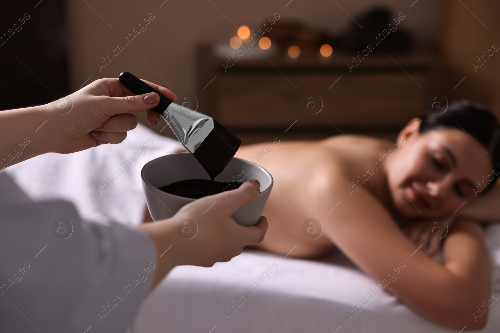 Photo of Spa worker with cosmetic product for chocolate body wrap near woman in salon, closeup. Selective focus