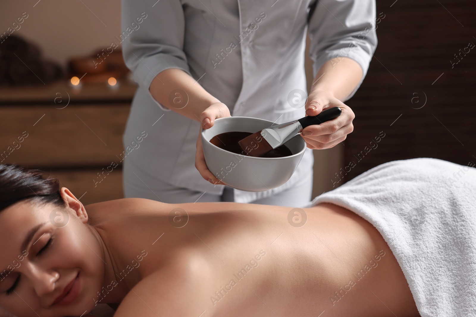 Photo of Spa worker with cosmetic product for chocolate body wrap near woman in salon, closeup