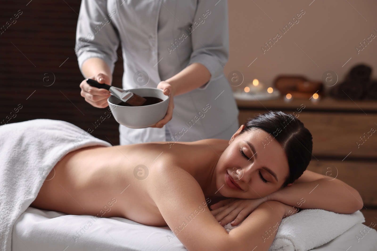 Photo of Spa worker with cosmetic product for chocolate body wrap near woman in salon, closeup