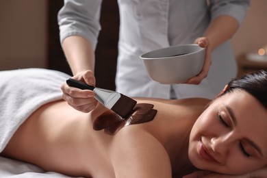 Photo of Chocolate body wrap. Spa worker applying mask onto woman's back in salon, closeup