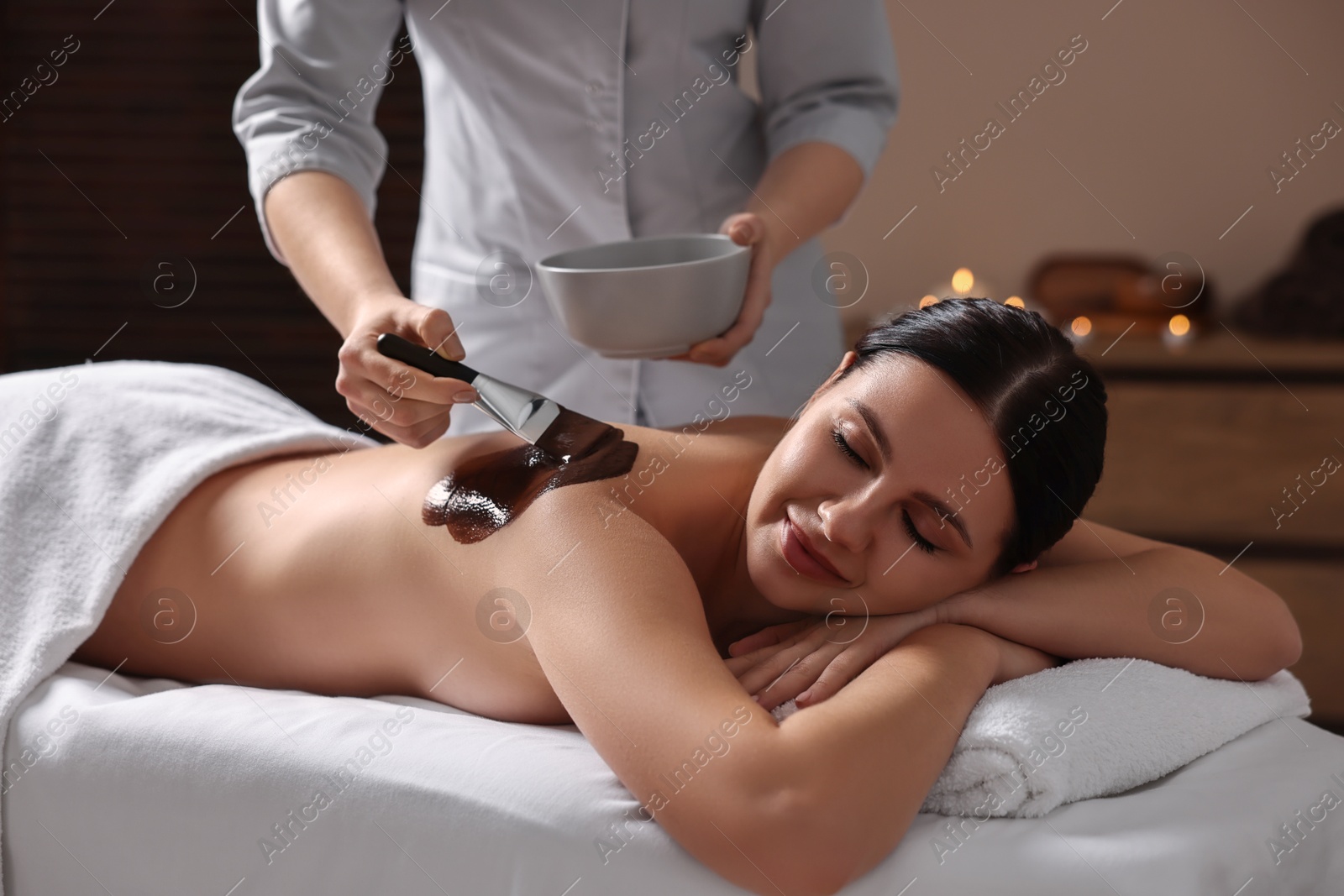 Photo of Chocolate body wrap. Spa worker applying mask onto woman's back in salon, closeup