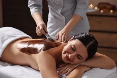 Photo of Chocolate body wrap. Spa worker applying mask onto woman's back in salon, closeup