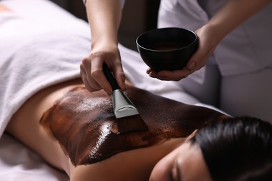 Photo of Chocolate body wrap. Spa worker applying mask onto woman's back in salon, closeup